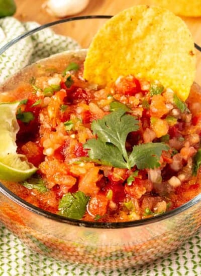 Tomato salsa in a bowl from the side with a chip and cilantro on top, a side of lime on the edge.