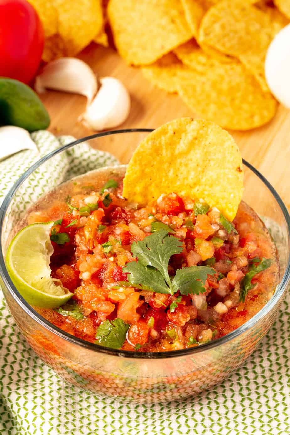 Blender salsa in a bowl with a chip from the side with more chips and fresh ingredients in the background.