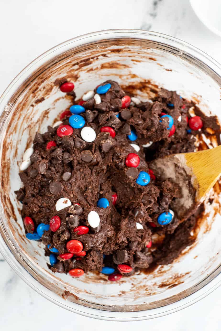 Folding red white and blue MMs into brownie cookie dough in a bowl from overhead with a wooden spoon.