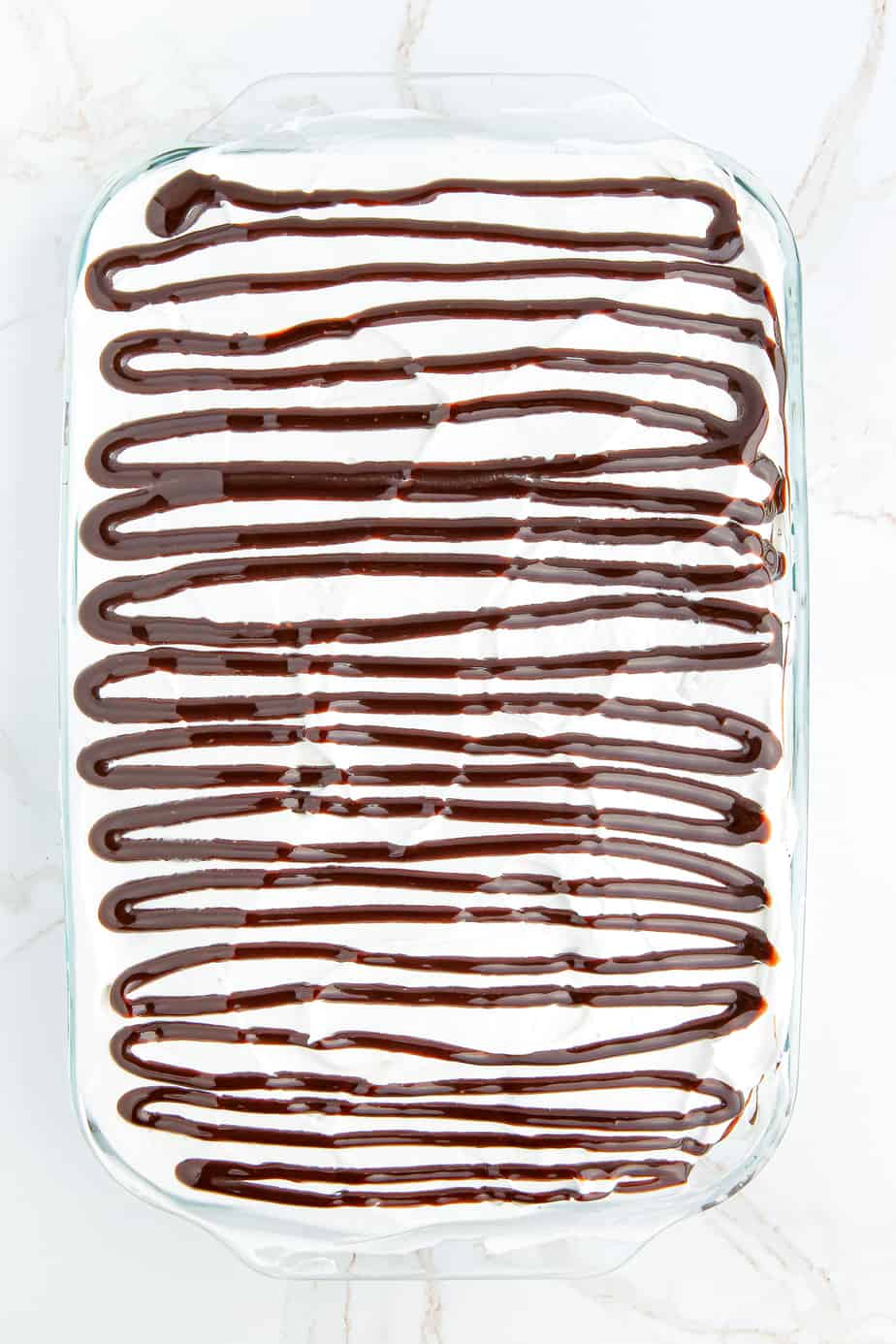 Chocolate sauce drizzled over whipped cream layer of an ice cream cake in a large glass rectangular baking dish.