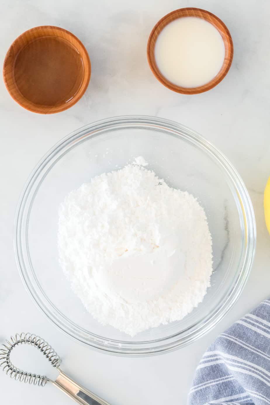 Powdered sugar, milk and lemon juice in bowls from overhead with a whisk on the counter nearby