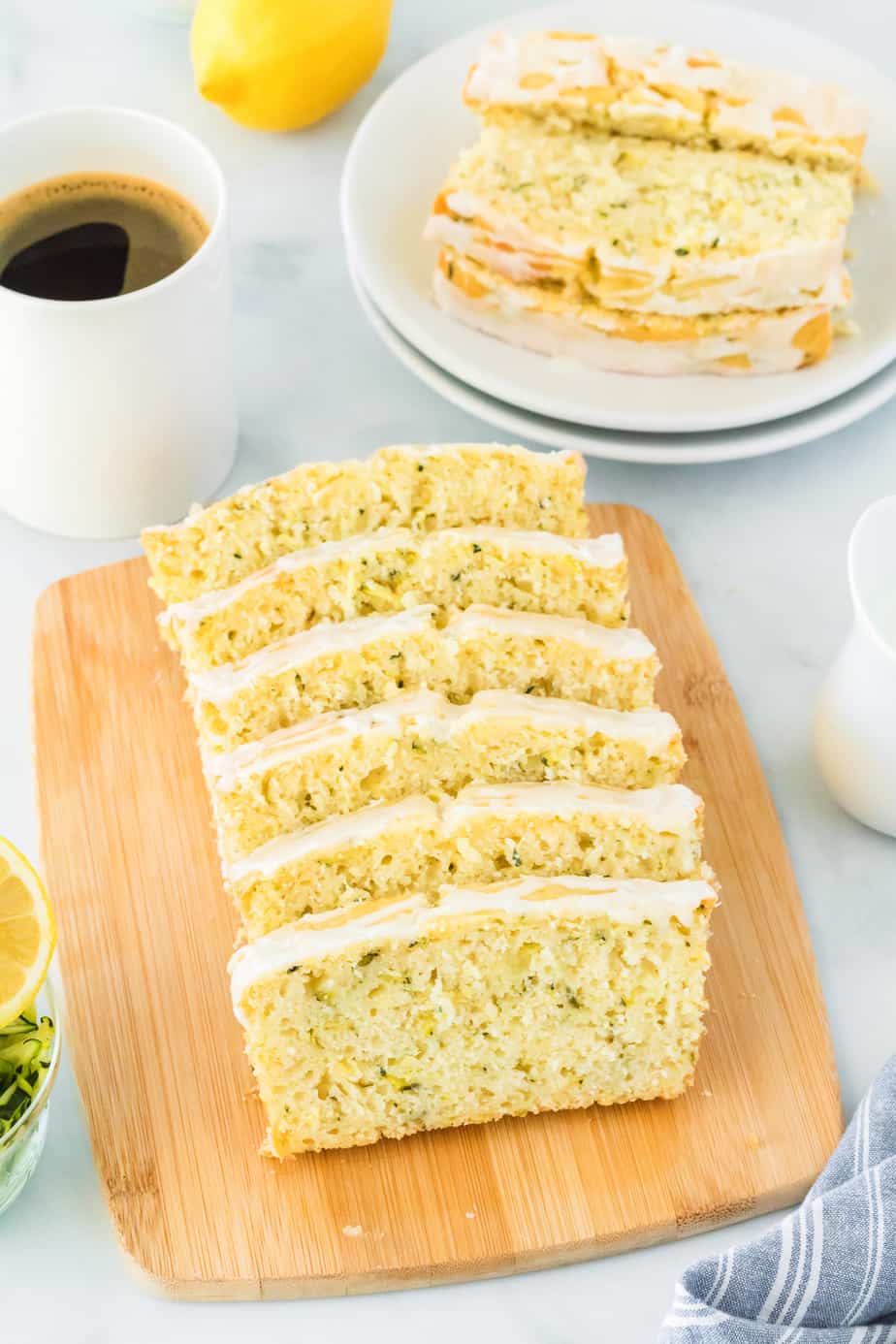Lemon zucchini cake loaf sliced on a platter with more slices on a plate behind the platter with a cup of coffee nearby