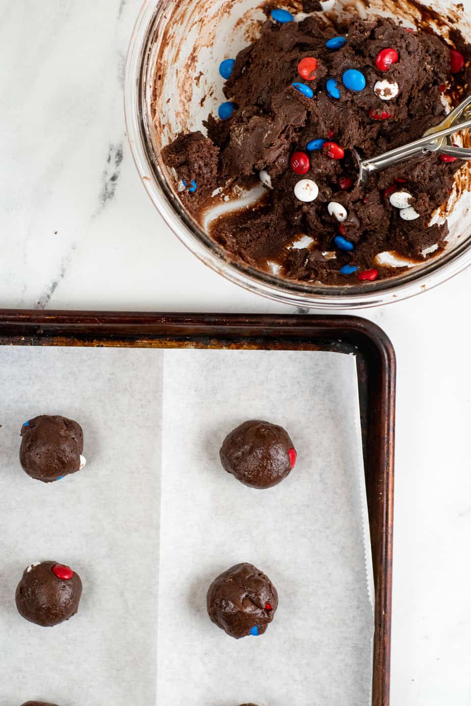 A baking sheet covered with spaced brownie mix cookie dough balls sits next to a bowl of brownie mix cookie dough with a scoop in it on a counter from above.