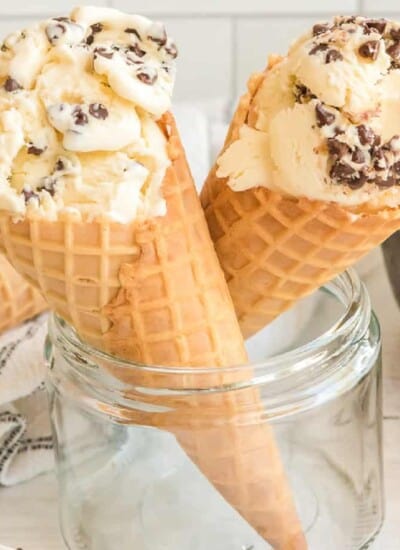 Two waffle cones resting in a glass container full of chocolate chip ice cream close up from the side.