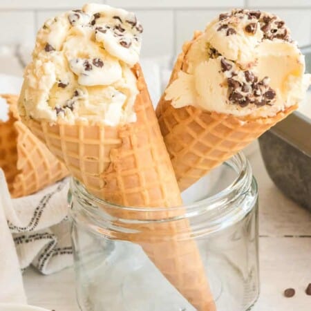 Two waffle cones resting in a glass container full of chocolate chip ice cream close up from the side.