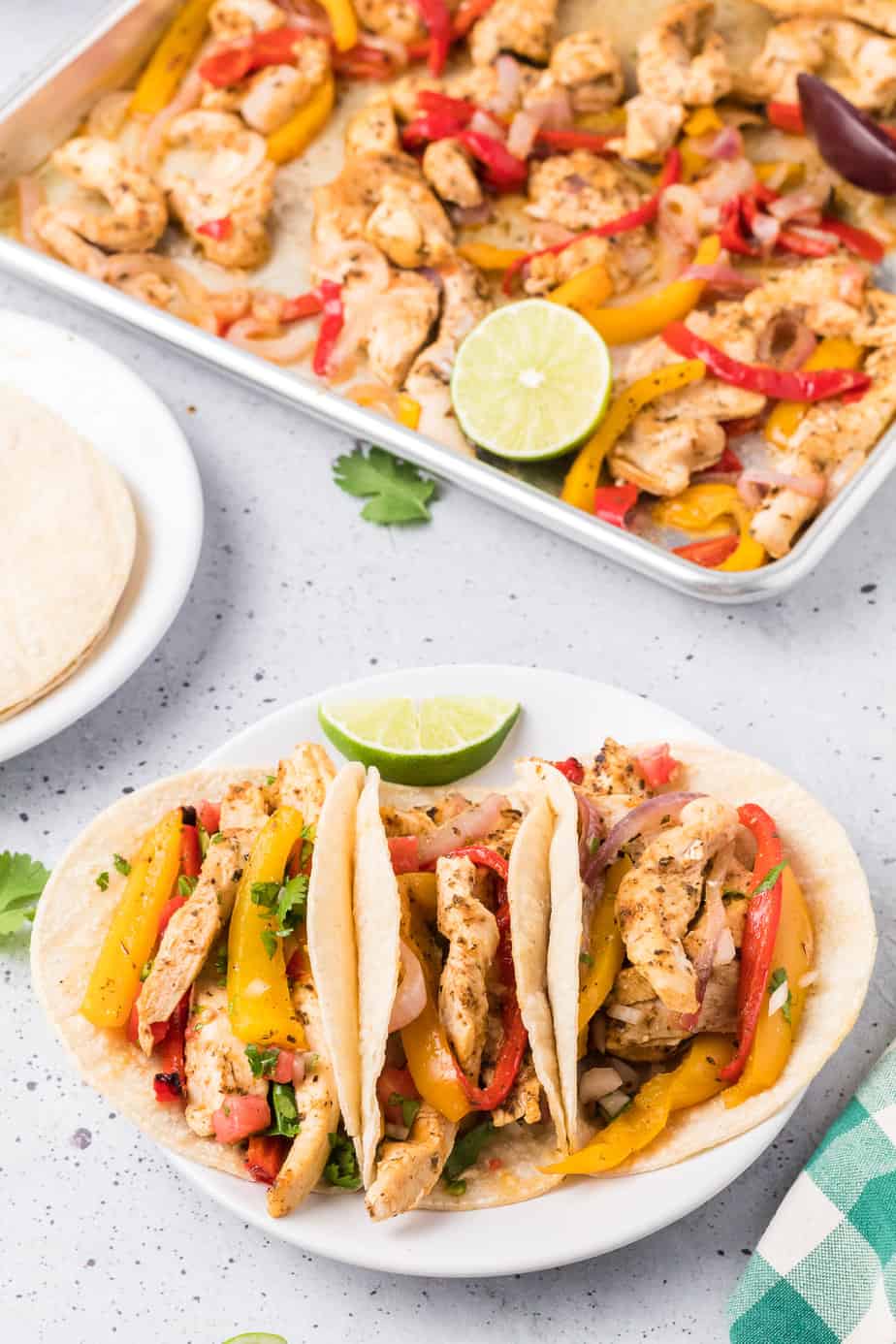 Chicken fajita tacos on a plate with a plate of tortillas and a sheet pan of chicken and vegetables in the background.
