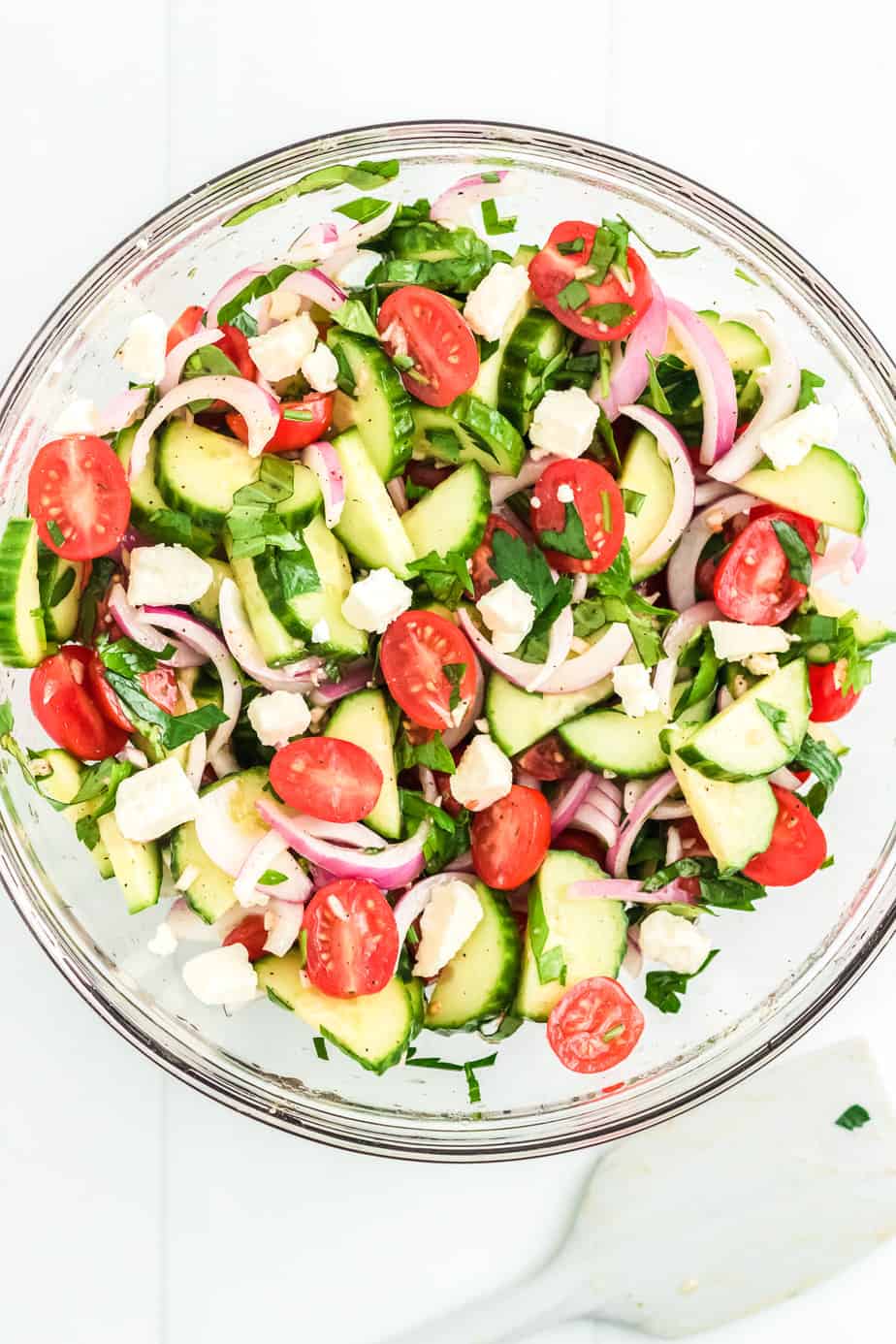 Mediterranean Cucumber tomato salad ingredients mixed together in a large mixing bowl from overhead tossed in dressing with a spatula next to the bowl.