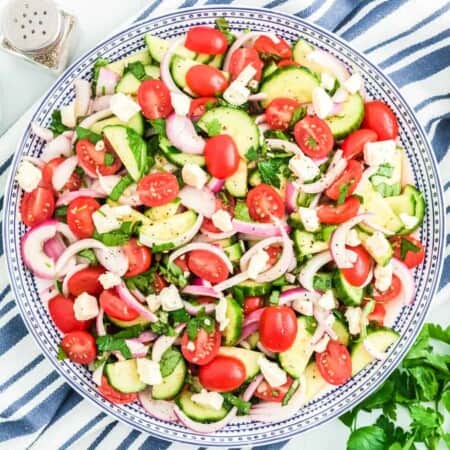 Cucumber tomato salad in a large serving bowl from overhead with a napkin with blue and white stripes, salt and pepper and fresh parsley near the bowl.