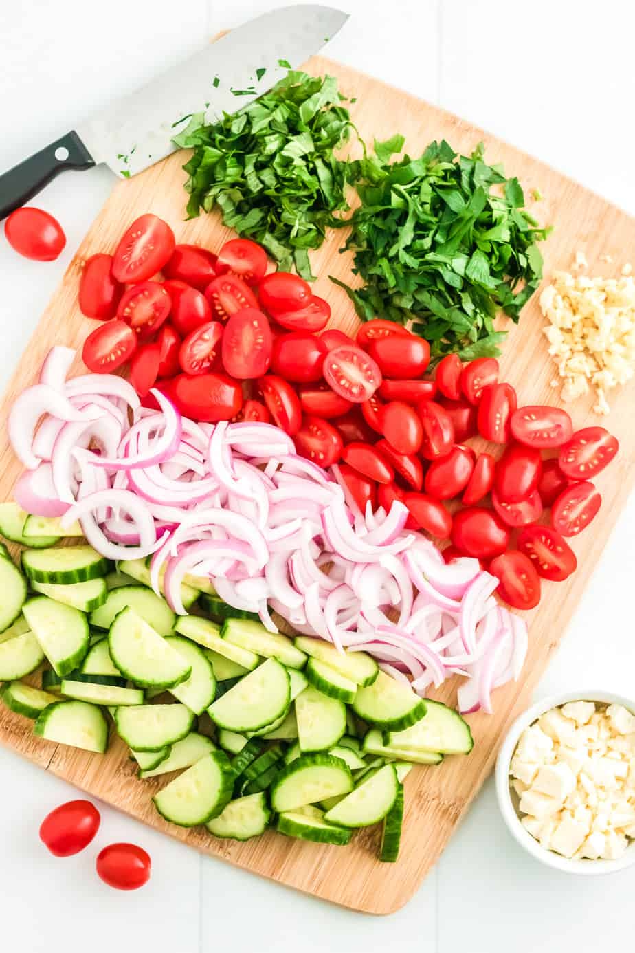 Cucumbers, red onions, tomatoes and fresh herbs chopped on a cutting board with a knife next to the cutting board from overhead.