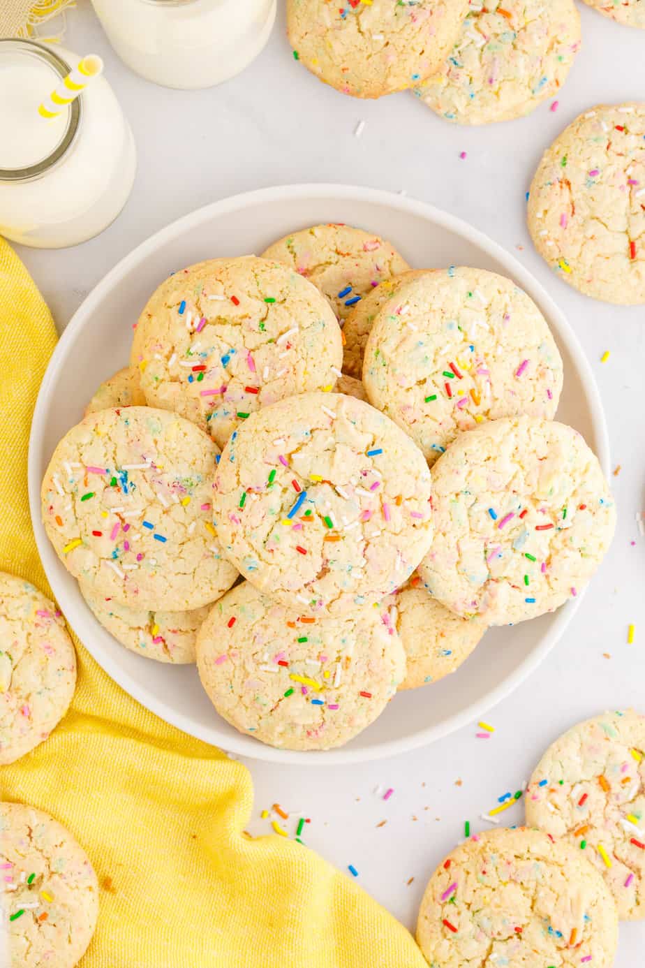 A plate of funfetti cookies with extra sprinkles on top from overhead on a table with milk and more cookies near the plate.