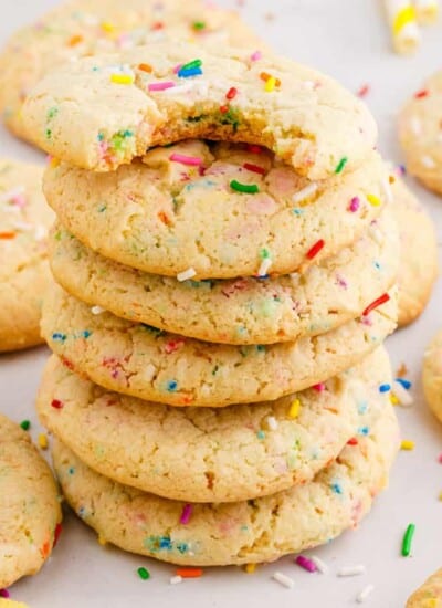 Square view of a stack of funfetti sprinkle cookies with the top cookie missing a bite and more cookies scattered next to the stack.