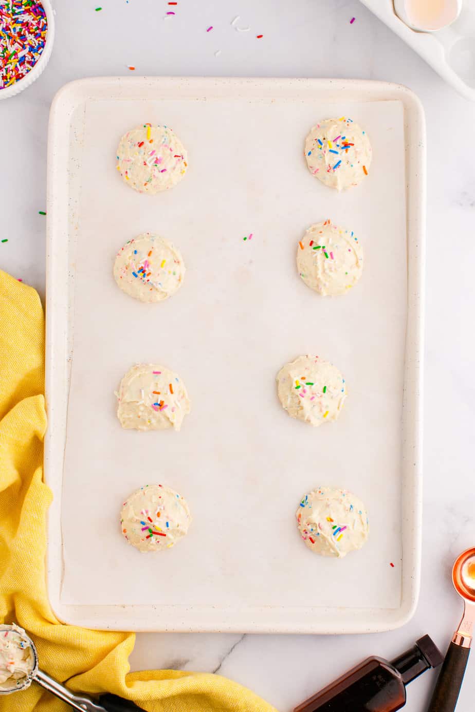 Funfetti cookie dough balls from above on a counter spaced out on a cookie sheet with extra sprinkles on top of each dough ball.