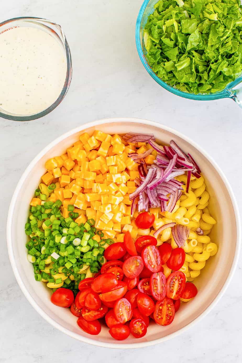 Chopped vegetables, cheese and cooked pasta in a bowl separated from overhead with a glass container of ranch and a bowl of chopped lettuce from overhead on the counter.