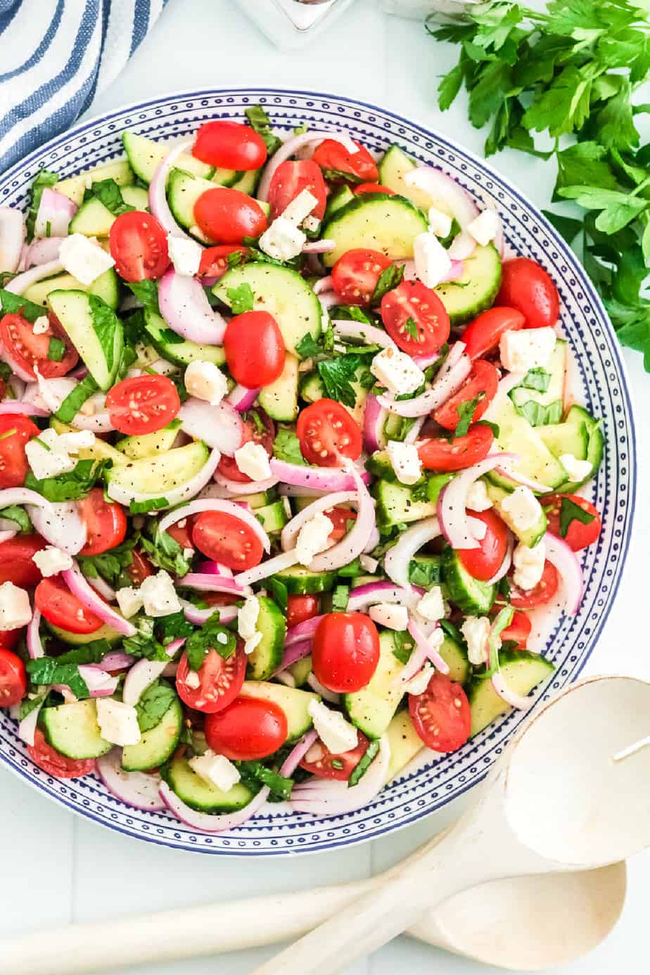 Mediterranean cucumber tomato salad in a large serving bowl from overhead with serving spoons an fresh herbs nearby.