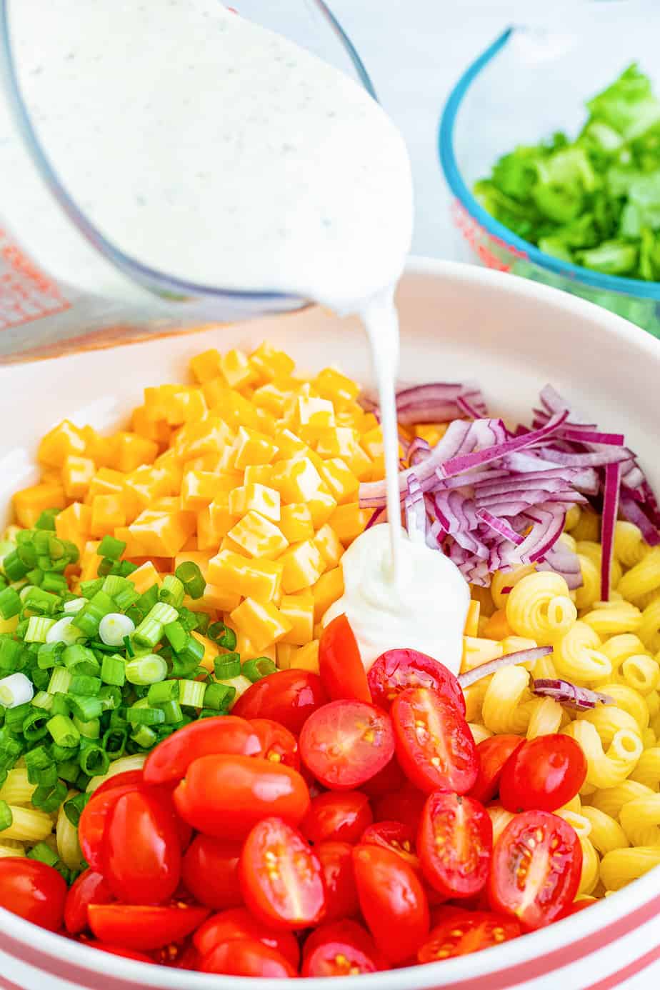 Pouring ranch dressing over a bowl full of cooked pasta, tomatoes, green onion, cheese and purple onion up close from the side.