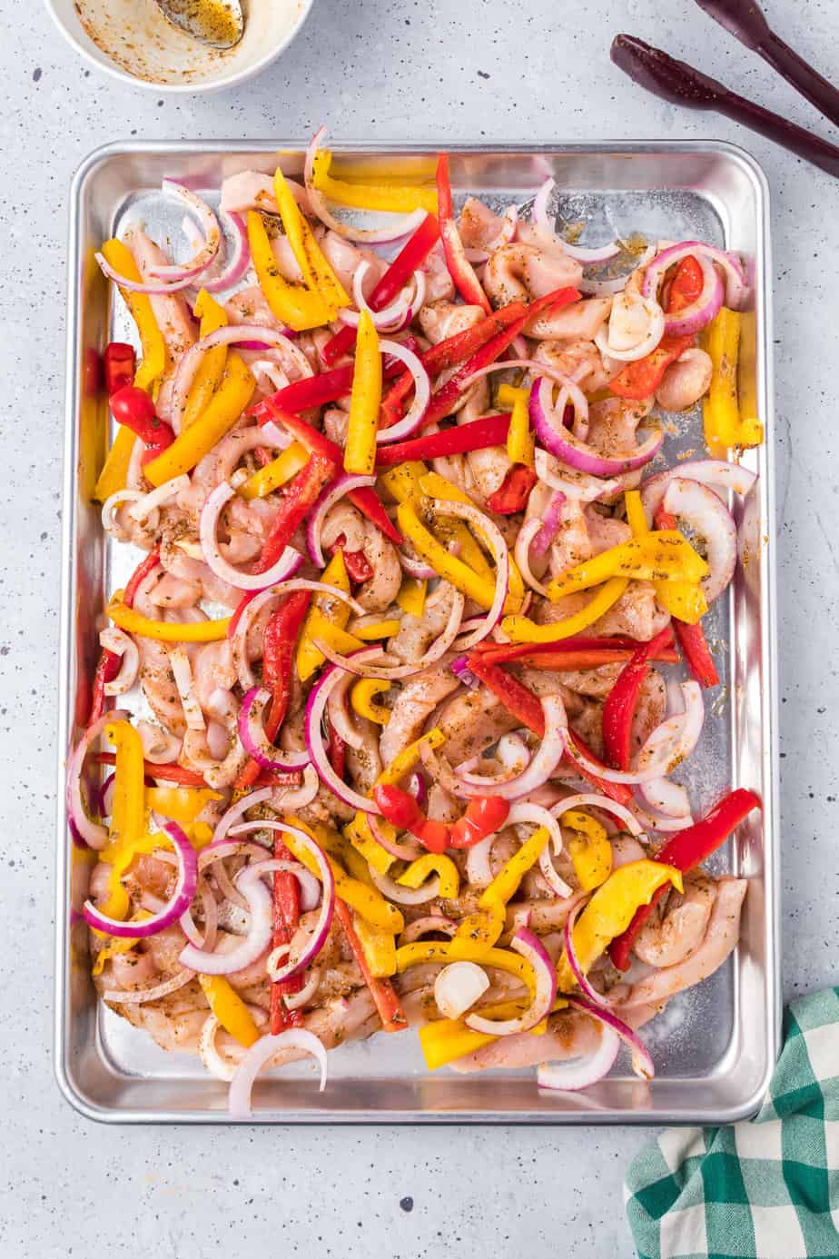 Raw chicken, bell peppers and onions cut into thin strips on a sheet pan tossed in oil and spices.