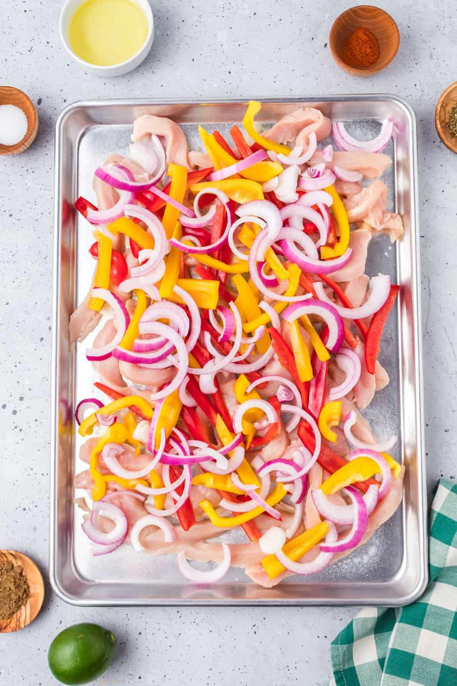 Thinly sliced raw bred and yellow bell peppers, purple onions and raw chicken on a sheet pan with other ingredients for the oil and spices in bowls nearby.