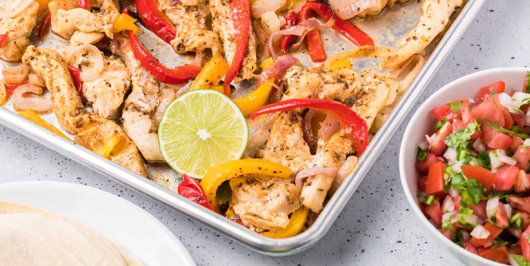 Wide view of chicken fajitas on a sheet pan with tortillas and pico de gallo on the table.