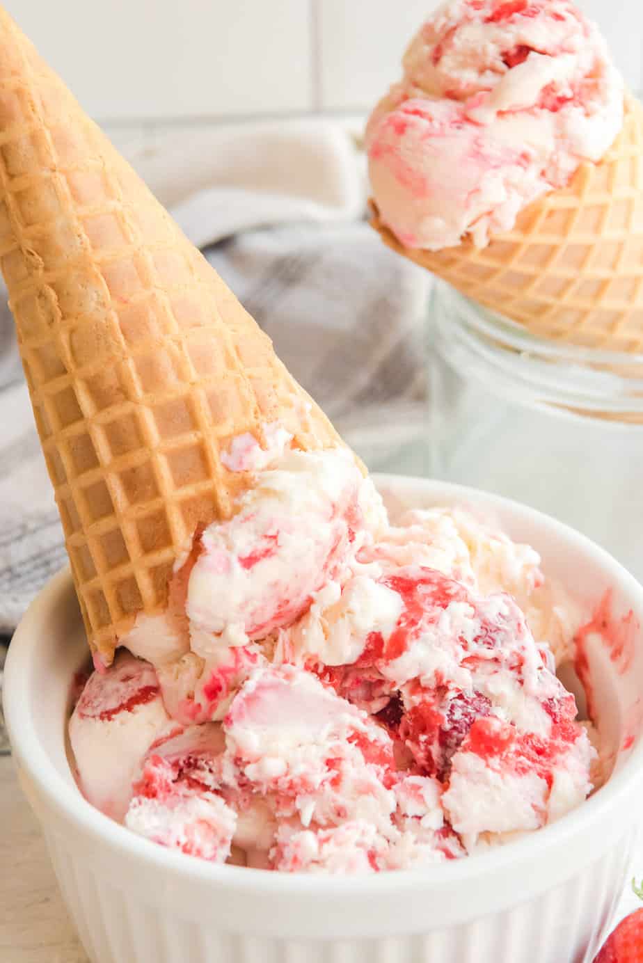 Strawberry swirl ice cream in a bowl with a cone on top and a strawberry ice cream filled cone in the background.