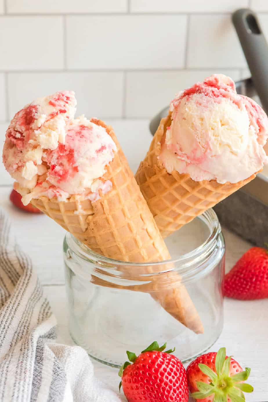 Two ice cream cones full of strawberry ice cream resting in a glass jar from the side with fresh strawberries nearby.