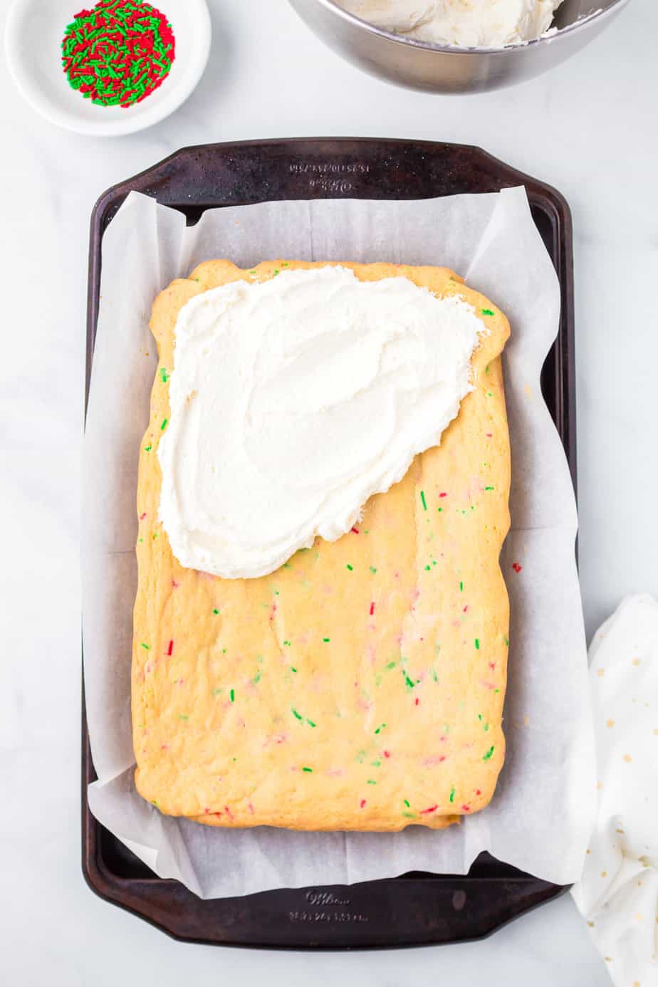 Adding frosting to the sugar cookie bars laying on parchment paper in a large pan from overhead.