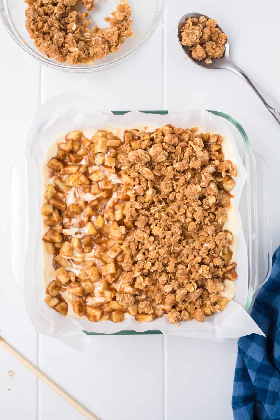 Adding crumble topping over apples and cheesecake layer in a square pan from overhead.