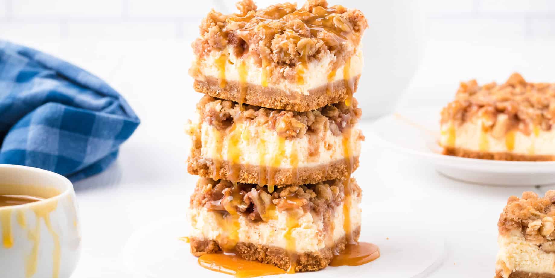 Wide short of stacked apple cheesecake bars drizzled with caramel with more bars in the background.