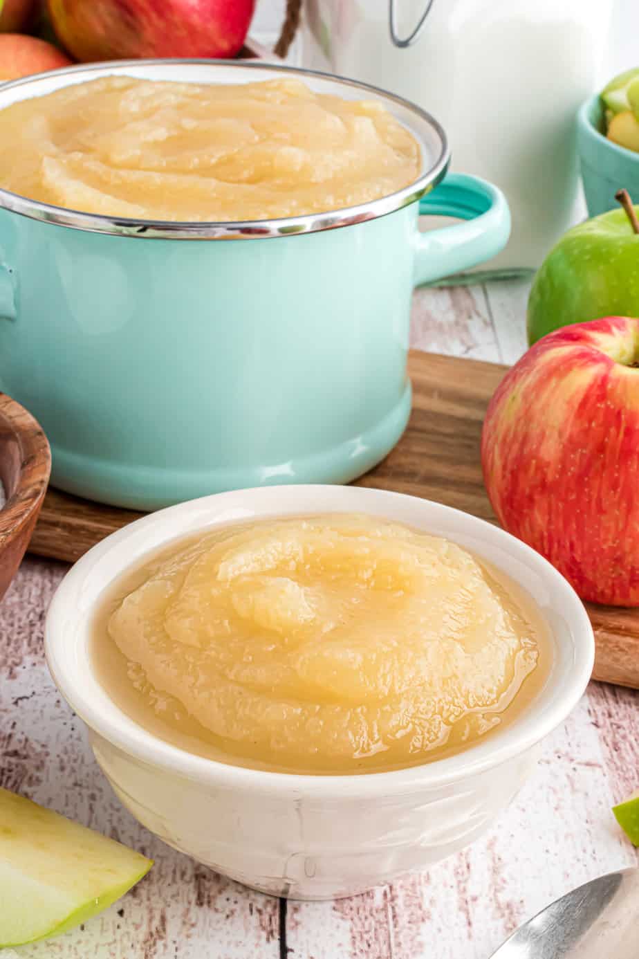 Applesauce in a bowl from the side with a fresh apple and a pot full of applesauce in the background.