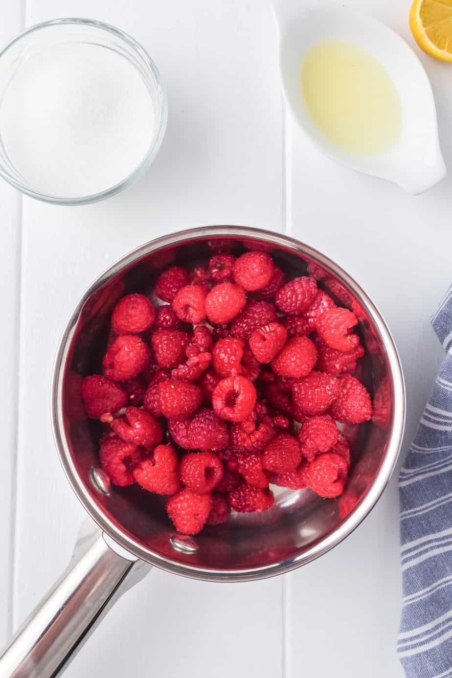 Raspberries in a pan with sugar and lemon juice nearby on the counter.