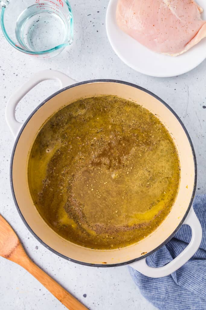 Spices on top of broth in a large pot with raw chicken and water on the counter in bowls nearby.