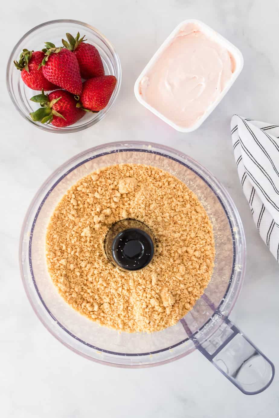 Golden Oreos in a food processor bowl with strawberry cream cheese and strawberries in a bowl nearby on the counter.