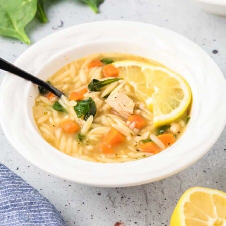 Bowl of soup from the side with chicken, orzo, spinach, carrot and slices of lemon.= and a spoon