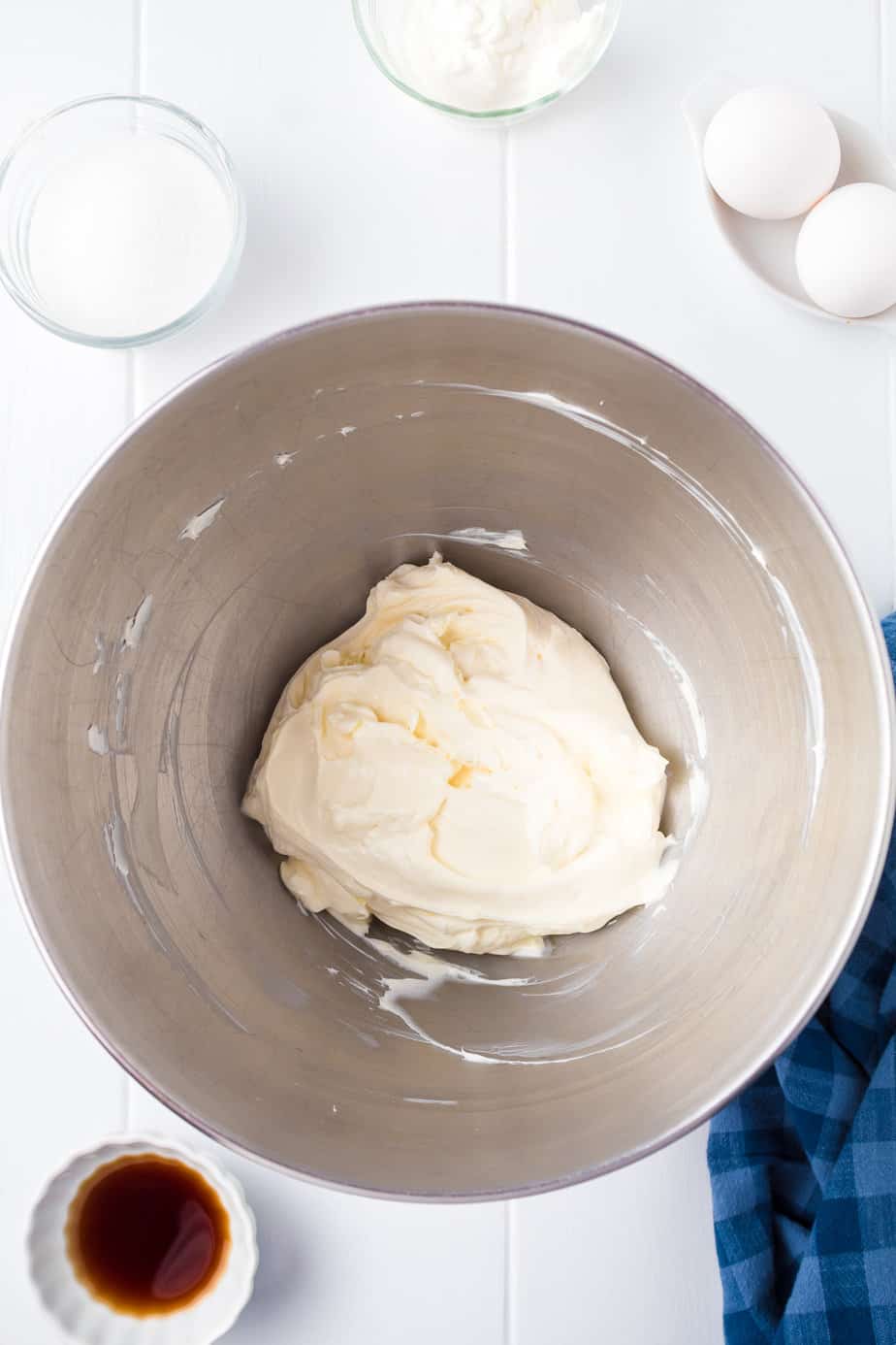 Softened cream cheese in a bowl to be mixed with bowls of vanilla, sugar and eggs nearby from overhead on a counter.