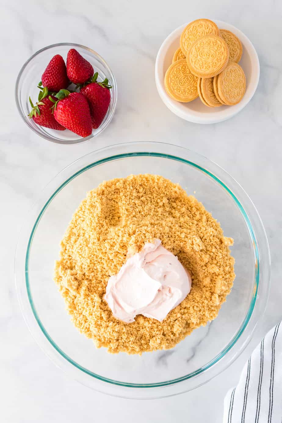 Mixing cream cheese into cookie crumbs in a bowl from overhead on the counter with strawberries and more cookies nearby.