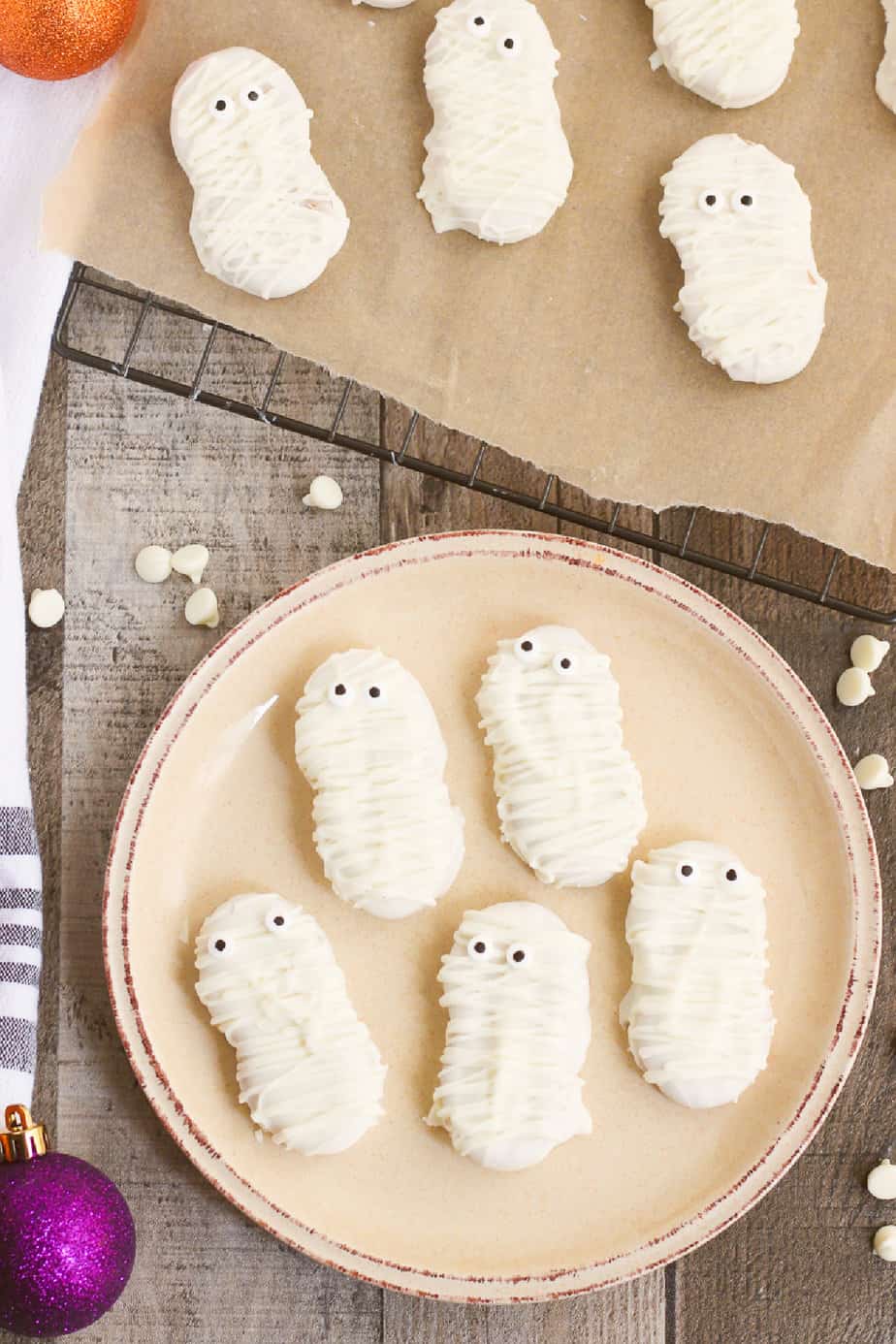 Cookies decorated to look like mummies with white chocolate on a plate with more on a tray on the table from overhead.