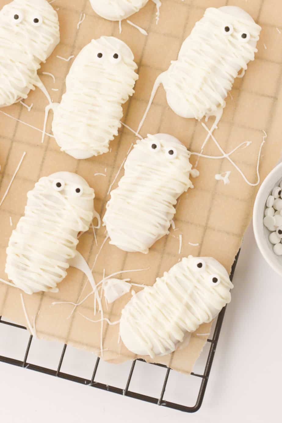 Nutter Butter cookies dipped in white chocolate and decorated to look like mummies with candy eyes cooling on a wire rack covered in parchment paper.