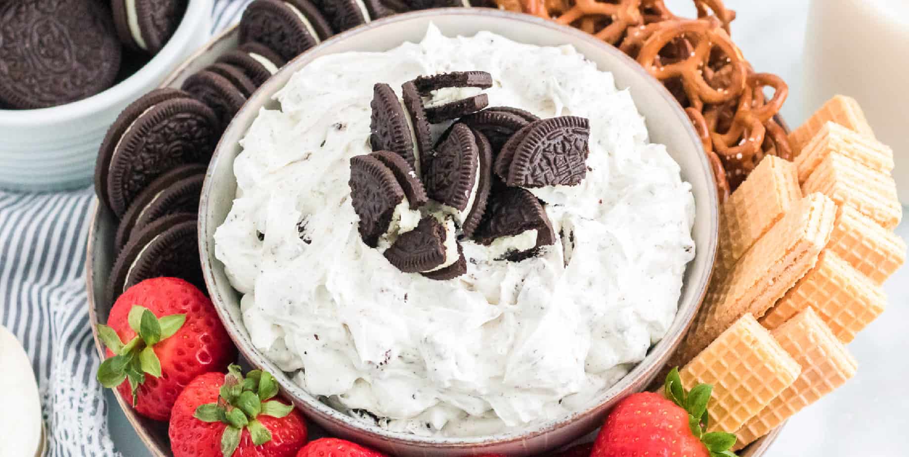 Wide view of a bowl of a dessert dip platter topped with Oreo pieces and surrounded by fruit, pretzels and cookies on the platter.