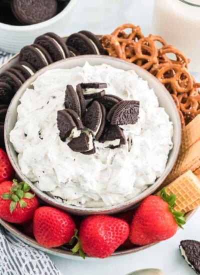Close up of a bowl of dessert dip topped with Oreo cookies on a round platter with more cookies, pretzels and strawberries on a counter.