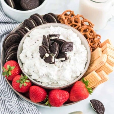 Close up of a bowl of dessert dip topped with Oreo cookies on a round platter with more cookies, pretzels and strawberries on a counter.