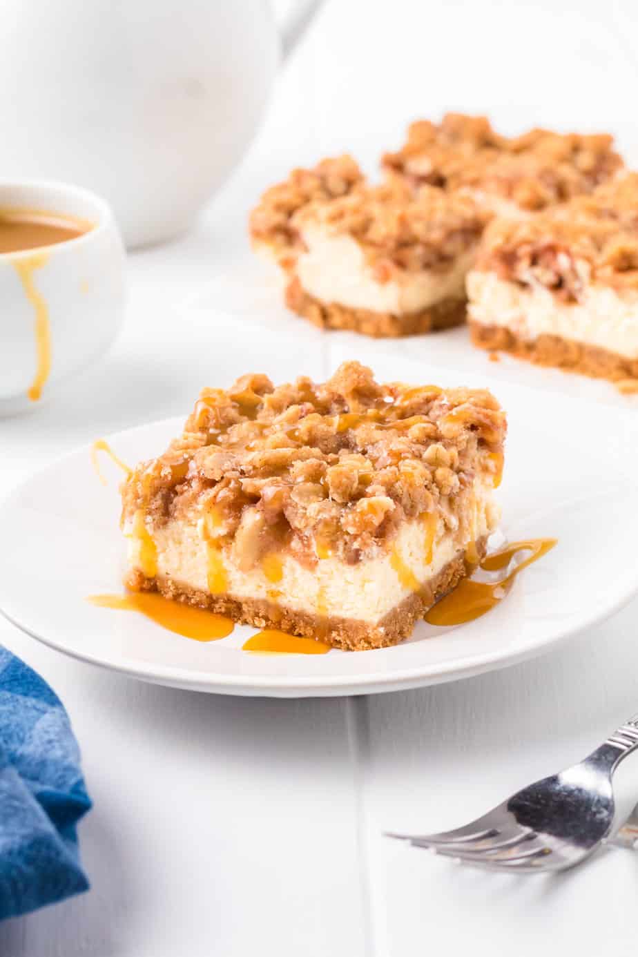 Plated apple cheesecake bar with a caramel drizzle from the side with more cheesecake bars and caramel in the background.