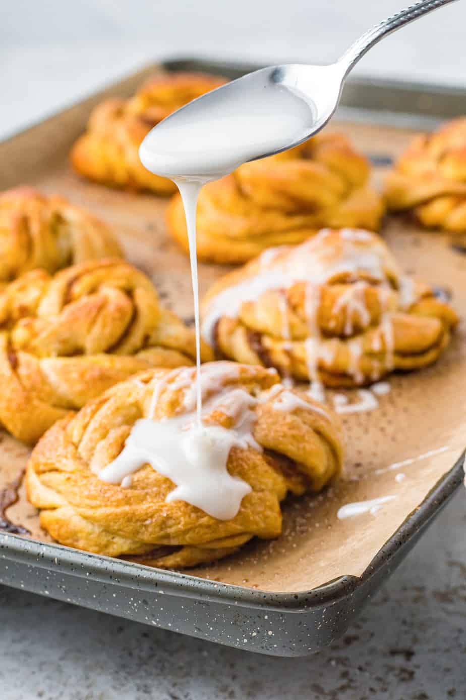 A spoon drizzles icing over a pumpkin crescent roll twist on a pan from the side.