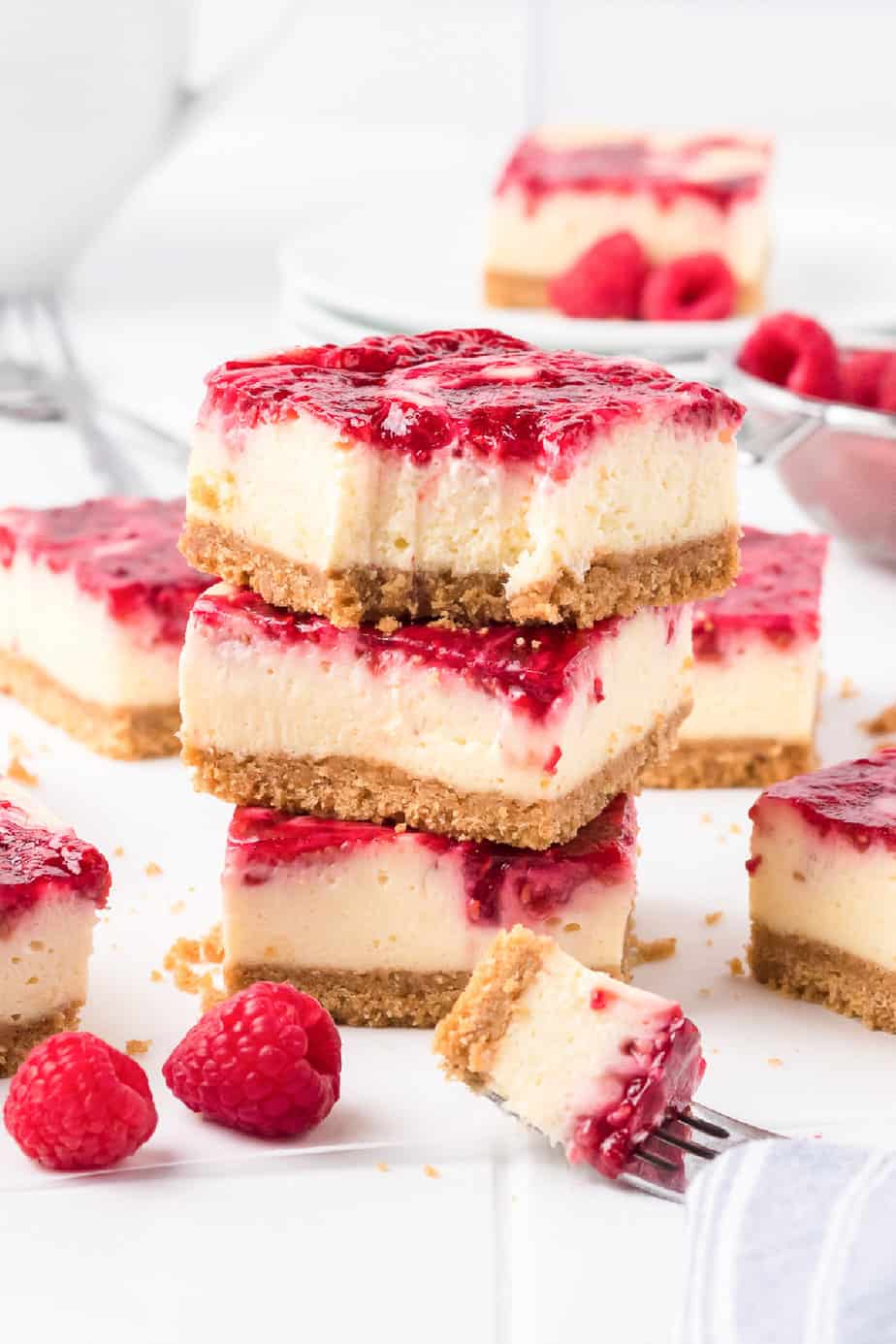 Raspberry swirl cheesecake bars stacked from the side with a bite missing from the top bar. More bars and fresh raspberries sit nearby on the counter, and a fork holds a bite of cheesecake.