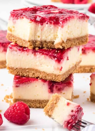 Stacked raspberry swirl cheesecake bars with the top bar missing a bite. Fresh raspberries and more cheesecake bars sit behind and nearby on the counter.