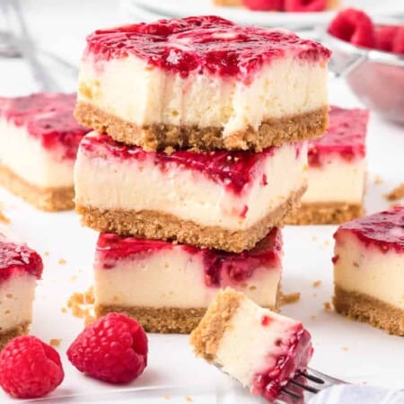 Stacked raspberry swirl cheesecake bars with the top bar missing a bite. Fresh raspberries and more cheesecake bars sit behind and nearby on the counter.
