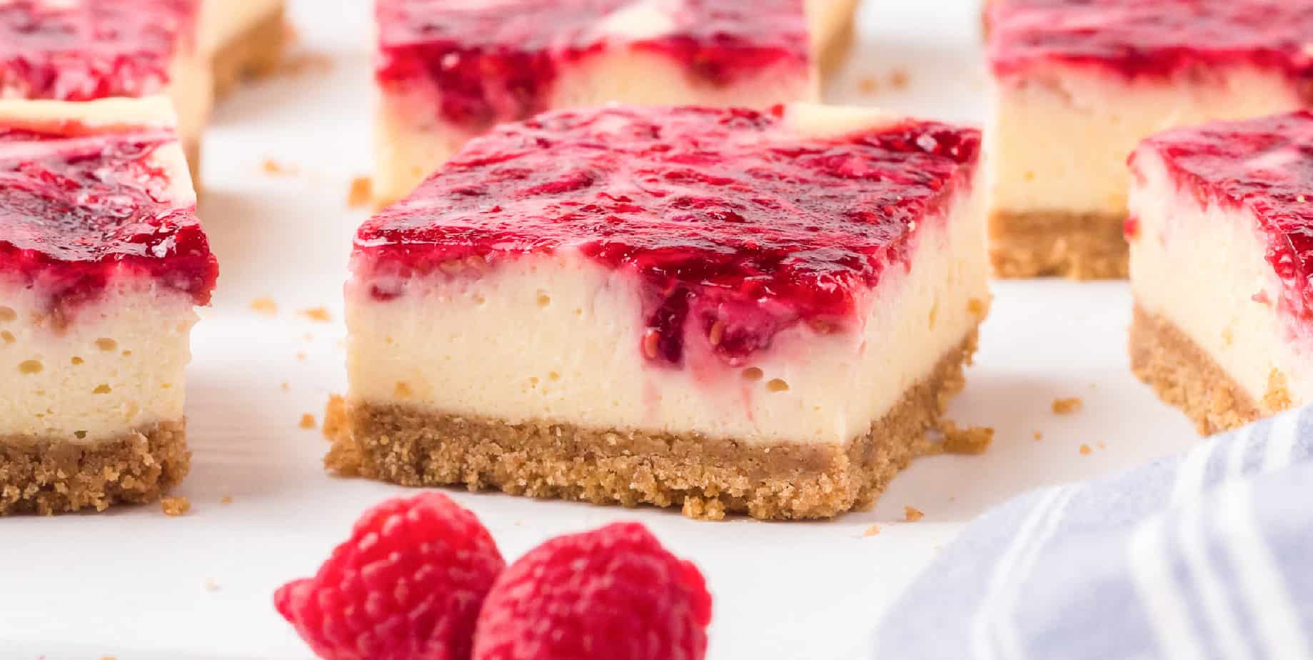 Wide close up of a raspberry cheesecake bar from the side with fresh raspberries and more bars nearby.