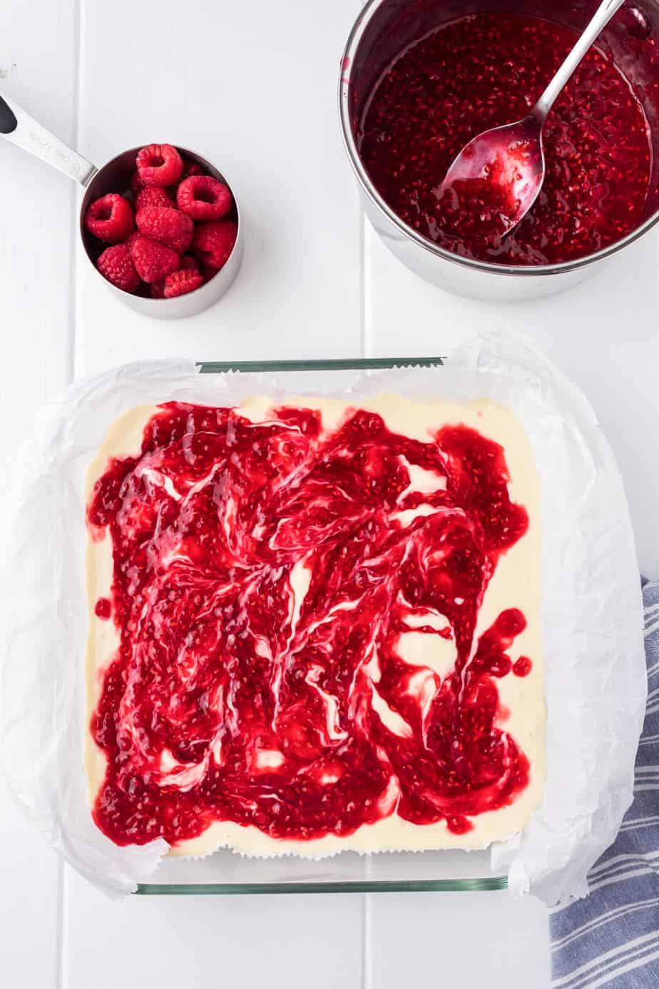 Raspberry sauce being swirled into cheesecake in a square pan from above with a raspberry saucepan nearby on the counter.