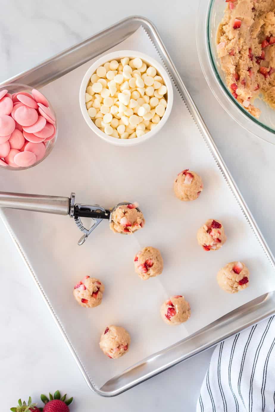 Using a scoop to portion strawberry truffles on a lines baking sheet with bowls of dough and white chocolate on the table nearby.