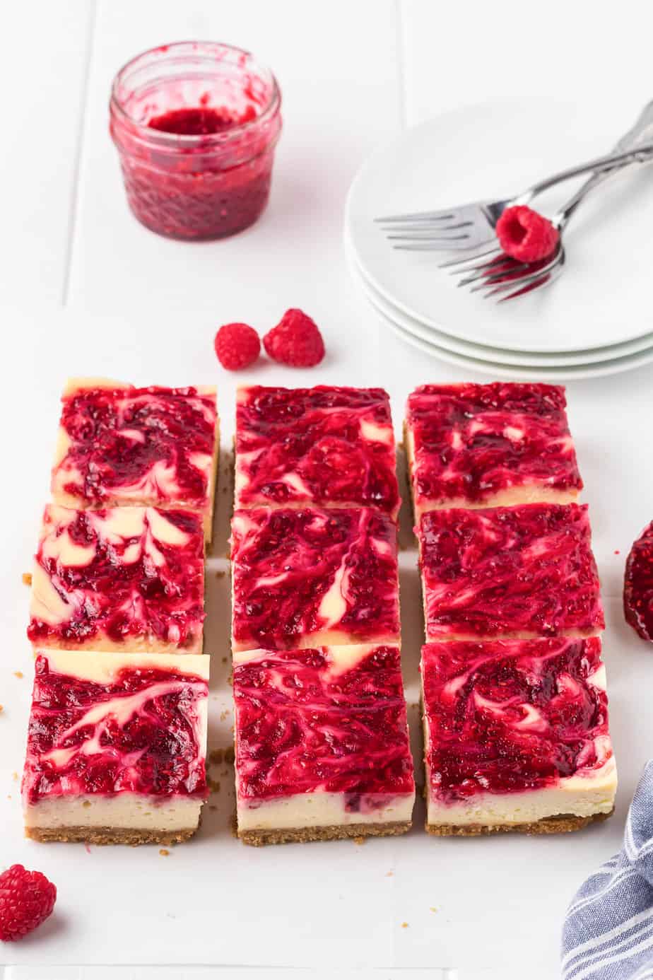 Sliced raspberry cheesecake bars on a counter with more fresh raspberries forks with plates, and raspberry sauce nearby.