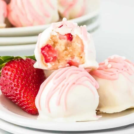 Close up of a strawberry shortcake truffle missing a bite showing strawberry bits inside on top of a few more truffles and a fresh strawberry on a plate.