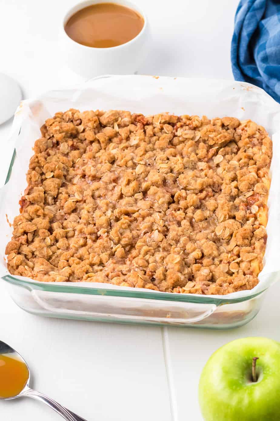 Apple crumb cheesecake bars after baking in a square pan from the side with an apple and caramel also on the table.